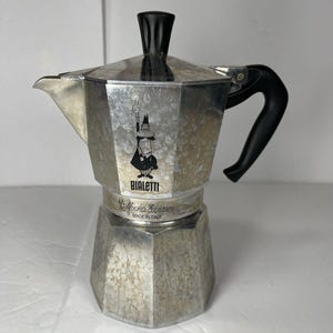 May include: A silver-colored Bialetti Moka Express stovetop espresso maker. The coffee pot has a black handle and a black knob on top. The Bialetti logo is visible on the front, with the words "Moka Express" and "Made in Italy".