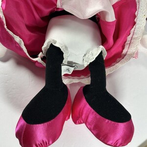 Op de afbeelding: Een pluche pop met een roze satijnen jurk en bijpassende schoenen. De pop heeft zwarte benen en voeten, met een wit ondergoed afgezet met kant. De jurk heeft een witte bovenlaag en een felroze rok.