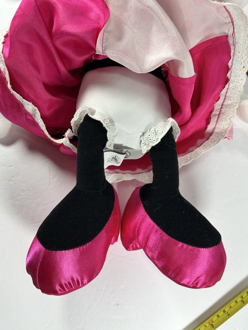Op de afbeelding: Een pluche pop met een roze satijnen jurk en bijpassende schoenen. De pop heeft zwarte benen en voeten, met een wit ondergoed afgezet met kant. De jurk heeft een witte bovenlaag en een felroze rok.