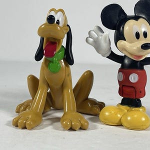 May include: Two plastic character figurines. One is a yellow dog with black ears, a red tongue, and a green collar. The other is a black, white, and red mouse character with large round ears, wearing red shorts with white buttons and yellow shoes.