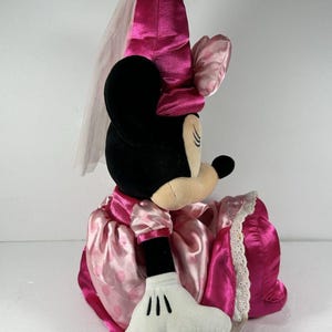 Op de afbeelding: Een Minnie Mouse pluche pop in een roze satijnen jurk met een witte kanten rand. De pop heeft een bijpassende roze strik en een sluier. De zwarte oren en het gezicht van de pop zijn zichtbaar, samen met een witte gehandschoende hand.