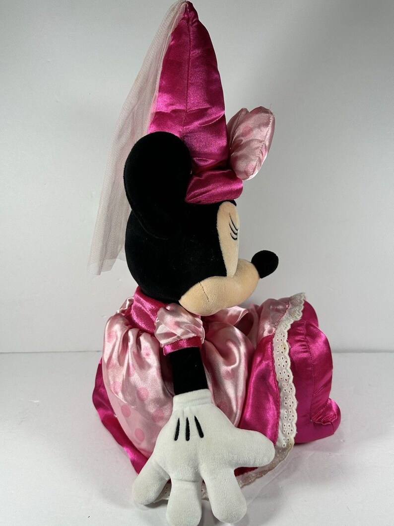Op de afbeelding: Een Minnie Mouse pluche pop in een roze satijnen jurk met een witte kanten rand. De pop heeft een bijpassende roze strik en een sluier. De zwarte oren en het gezicht van de pop zijn zichtbaar, samen met een witte gehandschoende hand.