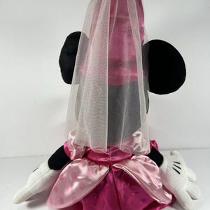 Op de afbeelding: Een pluche Minnie Mouse-pop met een roze satijnen jurk en een doorschijnende sluier. De pop heeft zwarte oren, witte handschoenen met zwarte details en een roze strik. De jurk heeft een gelaagd ontwerp met een donkerdere roze rok.