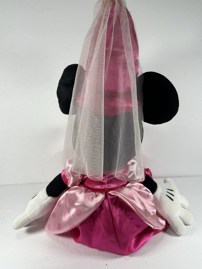 Op de afbeelding: Een pluche Minnie Mouse-pop met een roze satijnen jurk en een doorschijnende sluier. De pop heeft zwarte oren, witte handschoenen met zwarte details en een roze strik. De jurk heeft een gelaagd ontwerp met een donkerdere roze rok.