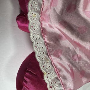 Op de afbeelding: Close-up van roze satijnen stof met witte kanten rand. De stof heeft een subtiele glans en een delicaat patroon van kleine, lichtere roze cirkels. De kanten rand is ingewikkeld gedetailleerd met een geschulpte rand.