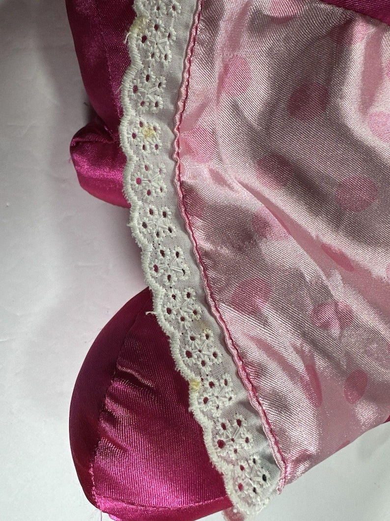Op de afbeelding: Close-up van roze satijnen stof met witte kanten rand. De stof heeft een subtiele glans en een delicaat patroon van kleine, lichtere roze cirkels. De kanten rand is ingewikkeld gedetailleerd met een geschulpte rand.