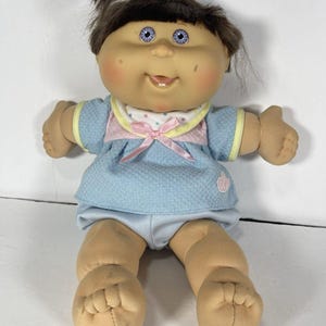 May include: A doll with brown hair tied in a ponytail, light skin, and blue eyes. It wears a textured light blue top with a pink bow, a white collar with colorful dots, and light blue shorts. The doll has a soft body and a vinyl head.