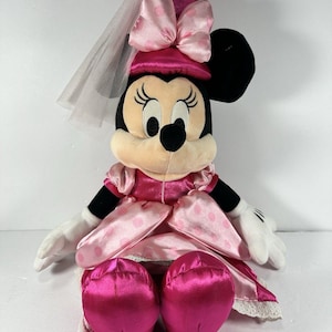 May include: A plush Minnie Mouse doll wearing a pink satin dress with white polka dots and a matching hat with a bow. The doll has a white veil and white gloves. The doll is sitting down.