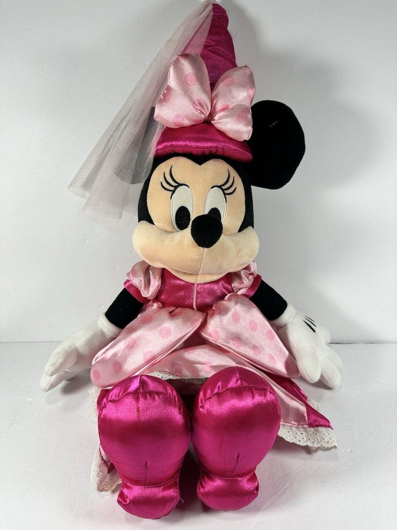 Op de afbeelding: Een pluche Minnie Mouse pop in een roze satijnen jurk met witte stippen en een bijpassende hoed met een strik. De pop heeft een witte sluier en witte handschoenen. De schoenen van de pop zijn felroze.