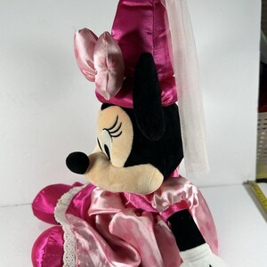 Op de afbeelding: Een pluche Minnie Mouse pop in een roze satijnen jurk met witte stippen en een bijpassend kapje. De pop heeft een witte sluier en een witte gehandschoende hand. Het gezicht van de pop is beige met zwarte ogen en een zwarte neus.