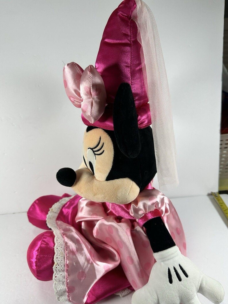 Op de afbeelding: Een pluche Minnie Mouse pop in een roze satijnen jurk met witte stippen en een bijpassend kapje. De pop heeft een witte sluier en een witte gehandschoende hand. Het gezicht van de pop is beige met zwarte ogen en een zwarte neus.