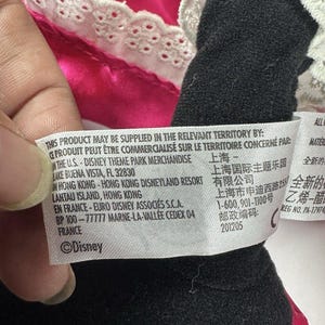 Op de afbeelding: Een close-up van een zwart pluche speelgoed met een roze jurk en witte kanten rand. Een label met het Disney-logo en tekst in meerdere talen is zichtbaar.