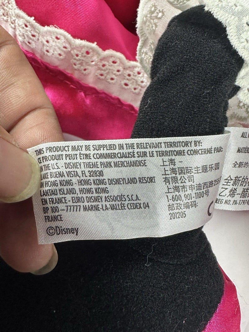 Op de afbeelding: Een close-up van een zwart pluche speelgoed met een roze jurk en witte kanten rand. Een label met het Disney-logo en tekst in meerdere talen is zichtbaar.
