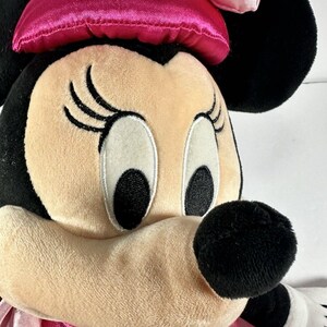 Op de afbeelding: Close-up van een Minnie Mouse knuffel. De knuffel heeft een roze strik, zwarte oren en een roze en witte polkadot jurk. Het gezicht is bruin met zwarte ogen, wimpers en een zwarte neus. Het speelgoed is gemaakt van zacht, pluche materiaal.