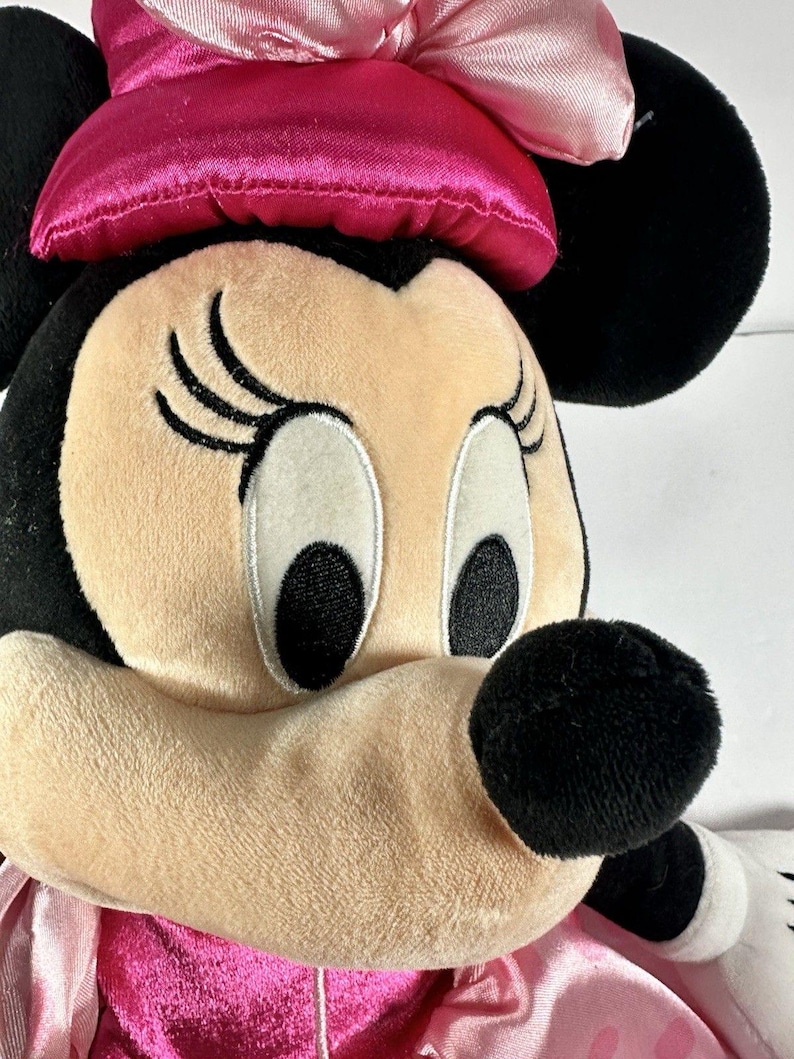 Op de afbeelding: Close-up van een Minnie Mouse knuffel. De knuffel heeft een roze strik, zwarte oren en een roze en witte polkadot jurk. Het gezicht is bruin met zwarte ogen, wimpers en een zwarte neus. Het speelgoed is gemaakt van zacht, pluche materiaal.