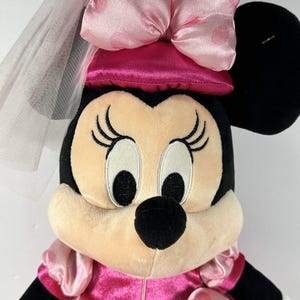 Op de afbeelding: Een pluche Minnie Mouse pop met een roze jurk met witte stippen, een bijpassende strik en een sluier. De pop heeft zwarte oren, een zwarte neus en grote ogen met lange wimpers. Een klassiek Disney-personage.