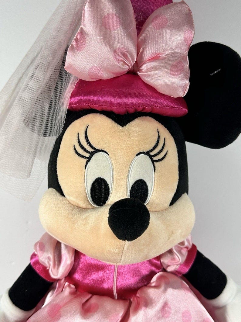 Op de afbeelding: Een pluche Minnie Mouse pop met een roze jurk met witte stippen, een bijpassende strik en een sluier. De pop heeft zwarte oren, een zwarte neus en grote ogen met lange wimpers. Een klassiek Disney-personage.