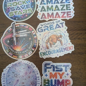 May include: Six decorative stickers with various designs. One is circular with space imagery and text 'GRACE ROCKY SAVE STARS'. Another says 'AMAZE' three times. A circular 'PROJECT HAIL MARY' sticker features a spacecraft. One shows a robot with 'WORDS OF GREAT ENCOURAGEMENT'. A circular sticker shows microscopic life with 'LIFE IS REASON STAR NOT DIE'. One says 'FIST MY BUMP'.