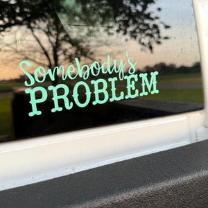 Könnte beinhalten: Ein türkisfarbener Vinylaufkleber mit den Worten "Somebody's PROBLEM" in einer verspielten Schriftart ist auf einer Fahrzeugscheibe angebracht. Der Hintergrund zeigt eine verschwommene Ansicht von Bäumen und einem Sonnenuntergang, die mit dem fetten Text kontrastiert.