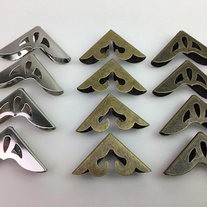 May include: An assortment of decorative metal corner protectors in silver and antique brass finishes. Each corner piece has a decorative cutout design. These are likely used for scrapbooking, crafting, or adding a vintage touch to projects.