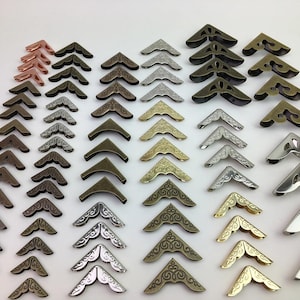 May include: A collection of metal corner protectors in various finishes including copper, bronze, silver, gold, and gunmetal. These V-shaped hardware pieces feature intricate embossed scrollwork patterns and smooth surfaces, arranged in rows.