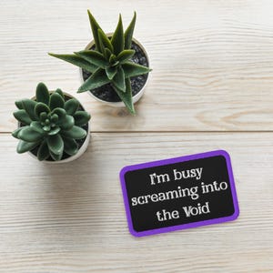 May include: Two small potted succulents with dark green leaves sit on a light wood surface. A rectangular sign with a purple border and the text "I'm busy screaming into the Void" is also present.