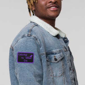 May include: A light blue denim jacket with a white sherpa collar. A purple patch on the jacket reads "Entering my Villain Era" with a silhouette of a witch's hat. The jacket has two front pockets and a button closure.