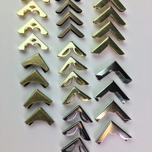 May include: Assortment of metal photo album corner protectors in various finishes, including gold, bronze, and silver. The protectors are in a classic V-shape, designed to secure and embellish the corners of photographs or albums. The image shows a variety of styles.
