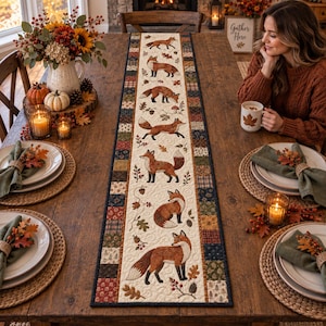 May include: A long rectangular table runner featuring a cream-coloured centre panel with a repeating pattern of illustrated foxes, acorns, and autumn leaves, bordered by a patchwork design of various dark-toned fabric squares in earthy colours.