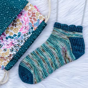 May include: A hand-knitted ankle sock in variegated teal, blue, and white yarn with a solid dark teal heel and toe, displayed on metal needles. Beside it is a fabric drawstring bag featuring a pattern of colourful tropical fish on white.