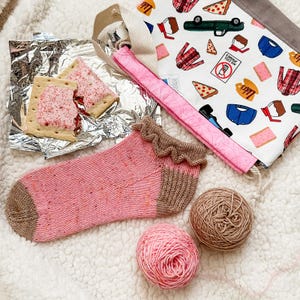 May include: A pink knitted sock with a frilled cuff and brown heel and toe sits next to two balls of yarn and a tote bag. The tote bag has a white background with colourful food and clothing graphics. Two frosted pastries are in foil.