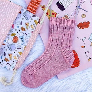 May include: A single, hand-knitted pink sock with ribbed detailing, displayed on a pink background. The sock is hanging from a metal hanger. The background includes a tote bag with a pattern of pizza slices, coffee cups, and autumn leaves.