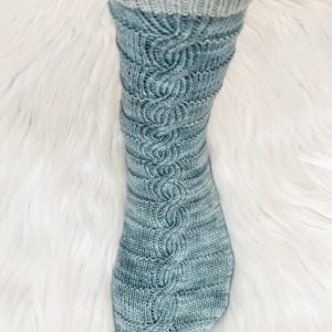 May include: A hand-knitted sock in shades of blue and grey, featuring a cable knit design. The cuff is a solid grey, transitioning to a textured blue pattern. The sock is displayed on a white, fluffy surface.