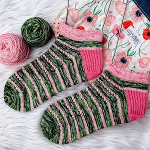 Puede incluir: Un par de calcetines tejidos a mano con un patrón de rayas verdes y rosas. El talón y el puño son de color rosa liso. Dos ovillos de lana, uno rosa y otro verde, están junto a los calcetines. Un tejido con estampado floral en el fondo.