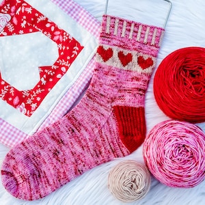 May include: A pink and red knitted sock with three red hearts near the top. The sock is displayed with red and pink yarn balls and a small quilted heart-shaped decoration. The sock has a red heel and toe.