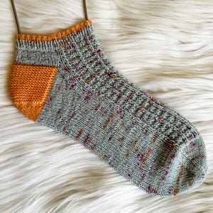 May include: A single, hand-knitted sock in shades of gray, orange, and purple. The heel and toe are a solid orange, while the main body features a speckled pattern. The cuff is trimmed with orange. The sock is displayed on a fluffy white surface.