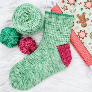 May include: A hand-knitted green and pink sock with matching yarn balls and a Christmas-themed fabric. The sock features a green and white speckled pattern on the leg and a solid pink heel and toe. The yarn balls are green and pink.