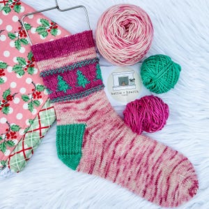 May include: A hand-knitted sock in shades of pink and green, with a Christmas tree pattern on the cuff. The sock is displayed with balls of yarn in pink and green, and a fabric with a holly berry pattern.