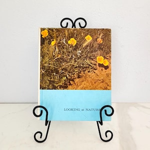 May include: A book titled "Looking at Nature" is displayed on a black metal stand. The book cover features a photograph of yellow flowers in a field. The stand has a scrolled design.
