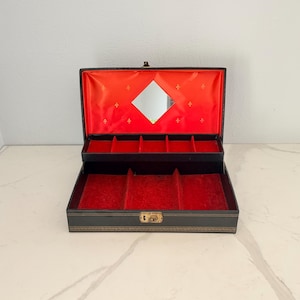 May include: Black jewelry box with a red velvet interior and a small mirror. The box is open, revealing compartments for storing jewelry. The exterior is black with gold-colored accents.