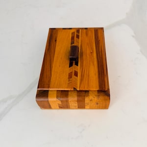 May include: A rectangular wooden box with a hinged lid. The box is made of various shades of brown wood, with a darker wood handle. The lid features a chevron pattern. The box is likely used for storage or display.