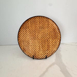 May include: A round, woven plate with a geometric pattern. The plate is made of natural-colored material and has a dark brown border. It is displayed on a black stand, suitable for serving food or as a decorative piece.