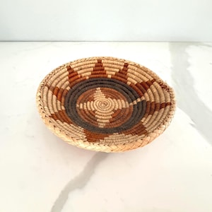 May include: A round woven basket with a spiral design in shades of brown, tan, and black. The basket has a shallow bowl shape and a textured surface. The design features a central spiral surrounded by concentric circles and star-like patterns.