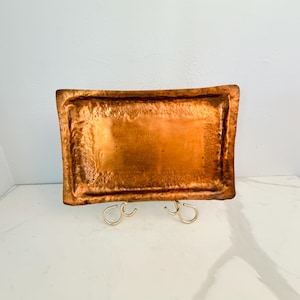 May include: A rectangular copper tray with a hammered, textured surface and raised edges. The tray features a warm, metallic orange-brown finish and is displayed on a small, gold-toned metal stand that supports the base of the tray.