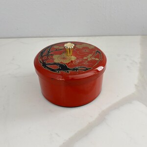 May include: A round, red lacquered box with a lid featuring a floral design in gold and black. The lid has a gold-colored knob. The box is sitting on a white surface.