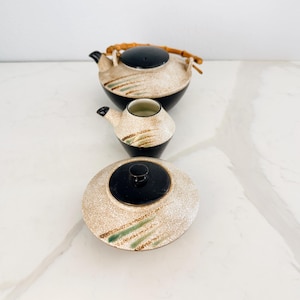 Könnte beinhalten: Eine weiße Keramik-Tasse mit schwarzem Henkel und dem Text "But First, Coffee" in fetter, schwarzer Schrift. Die Tasse steht vor einem verschwommenen Hintergrund, der eine gemütliche Umgebung suggeriert.