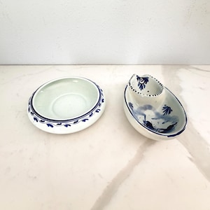 Może przedstawiać: Dwa białe ceramiczne naczynia do serwowania z niebieskimi kwiatowymi wzorami. Jedno jest owalne z podniesioną częścią, drugie to okrągła miska. Naczynia te służą prawdopodobnie do serwowania sosów lub małych porcji.