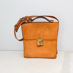 May include: A vintage, rectangular leather satchel in a warm, tan color. It features a top handle and an adjustable shoulder strap. The bag has a brass clasp closure and brass hardware. The satchel is approximately 12 inches tall, 10 inches wide, and 4 inches deep.