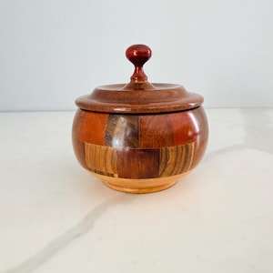 May include: A handcrafted wooden jar with a lid. The jar is made of various wood tones, including reddish-brown and light brown. The lid has a rounded shape with a small, dark red knob. The jar sits on a light-colored surface.