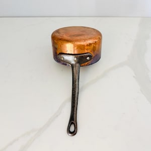 May include: A round copper saucepan featuring a dark, weathered metal handle attached with three visible rivets. The exterior of the copper pan shows a warm, metallic finish with signs of natural oxidation and discoloration along the sides.